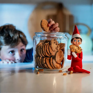 Poseable Elf on the Shelf doll in red suit holding chocolate chip cookie beside cookie jar