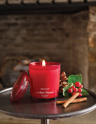 Aromatique red glass jar candle burning beside cinnamon sticks, pine cone, and holly berries