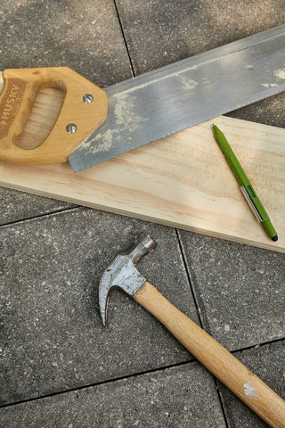 Olive green Fw Tooling Pen resting on a wooden plank alongside a handsaw and hammer