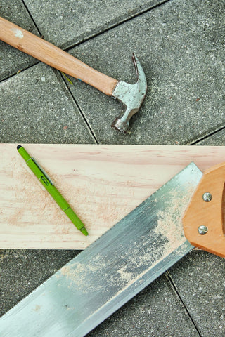 Olive green Fw tooling pen resting on a wooden plank alongside a handsaw and hammer