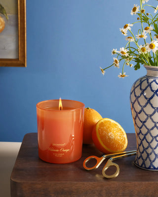 Aromatique Valencia Orange 10oz orange glass candle, lit, beside fresh oranges and daisy vase